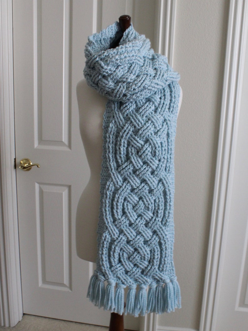 May include: A light blue knitted scarf with a braided pattern and tassels. The scarf is displayed on a white mannequin.
