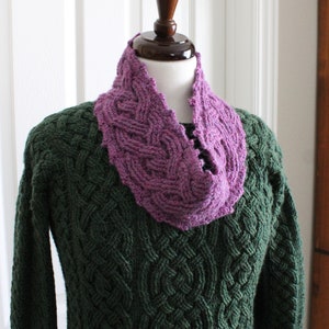 May include: A purple knitted cowl with a cable knit pattern. The cowl is worn on a mannequin with a green sweater.