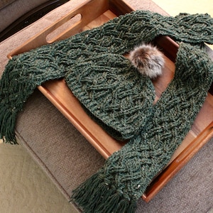 May include: A green knitted scarf with a braided pattern and fringe tassels. The scarf is displayed on a wooden tray with a faux fur pom-pom.