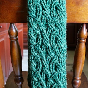May include: A dark green knitted scarf with a cable knit pattern. The scarf is draped over the back of a wooden chair.