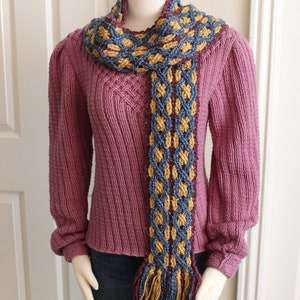 May include: A knitted scarf with a braided pattern in shades of blue, gold, and burgundy. The scarf is draped over a mannequin wearing a pink sweater and blue jeans.