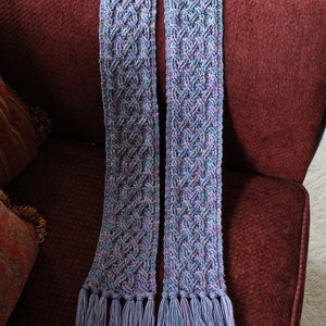 May include: A long, purple and blue knitted scarf with fringe tassels. The scarf is made with a cable knit pattern.