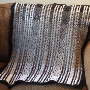 Può includere: Una coperta in crochet nera, bianca e grigia con un motivo a treccia.