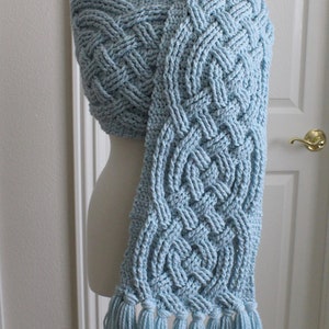 May include: A light blue crocheted scarf with a Celtic knot pattern and tassels. The scarf is draped over a mannequin.