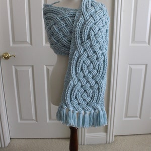 May include: A light blue crocheted scarf with a Celtic knot pattern and fringe. The scarf is displayed on a wooden mannequin.