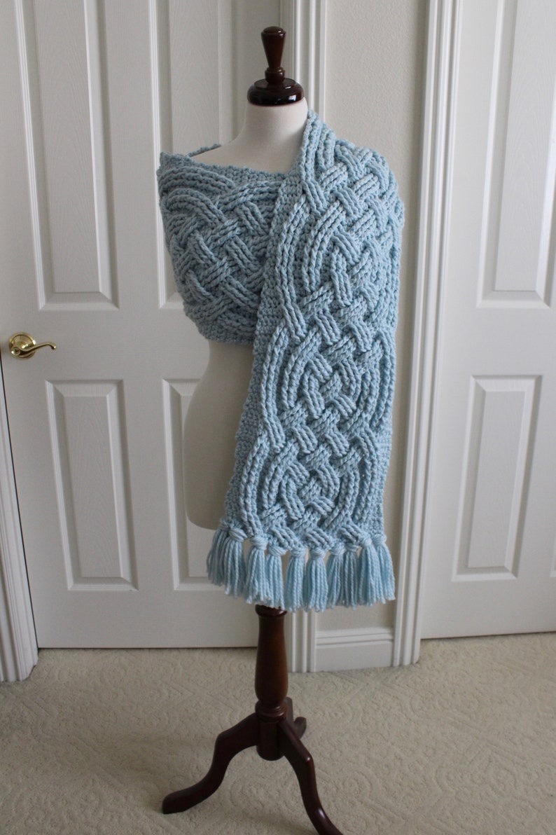 May include: A light blue crocheted scarf with a Celtic knot pattern and fringe. The scarf is displayed on a wooden mannequin.