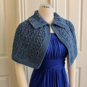 May include: A blue knitted cape with a Celtic knot pattern. The cape has a collar and a button fastening. It is worn over a blue dress.