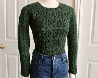 May include: A green cable knit jumper with a round neckline. The jumper is on a mannequin and is paired with blue jeans.