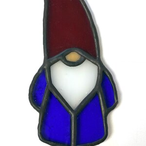 May include: Stained glass garden gnome with a red hat, white beard, and blue jacket. The gnome is mounted on a black metal stake for easy placement in a garden or flower bed. A charming and colorful outdoor decoration.