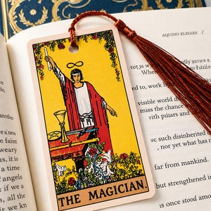 May include: A rectangular bookmark featuring The Magician tarot card illustration with a yellow background, a figure in a red robe, and a table with ritual items. The bookmark has a hole at the top with a long, dark red silk tassel attached.