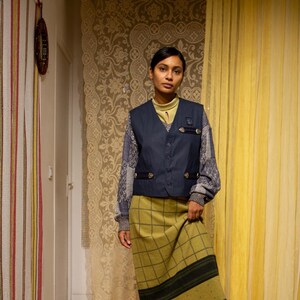 May include: A woman in a navy blue waistcoat with gold buttons, a patterned long-sleeved top, a green plaid skirt with a fringed hem, and brown knee-high boots. The setting is a room with patterned curtains and a decorative rug.