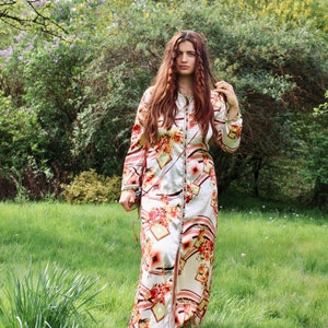 May include: A woman wearing a long, white dress with a floral pattern and a brown belt. The dress has a long, flowing skirt and a fitted bodice. The woman is standing in a grassy field with trees in the background.