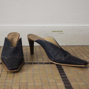 Peut inclure: Une paire de mules en jean bleu foncé avec un bout pointu et un talon haut. L'avant de la chaussure présente un empiècement en cuir texturé façon crocodile avec une découpe en V. La semelle intérieure est beige et le talon est en bois brun.