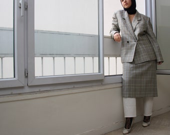 Può includere: Una donna indossa un blazer e una gonna a quadri grigi con un foulard nero. È in piedi vicino a una finestra con una ringhiera bianca.