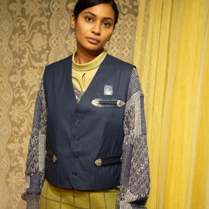 May include: A navy blue waistcoat with silver-toned details and a small square badge. It's worn over a patterned jumper and a pale green roll neck. The outfit is completed with a green plaid skirt.