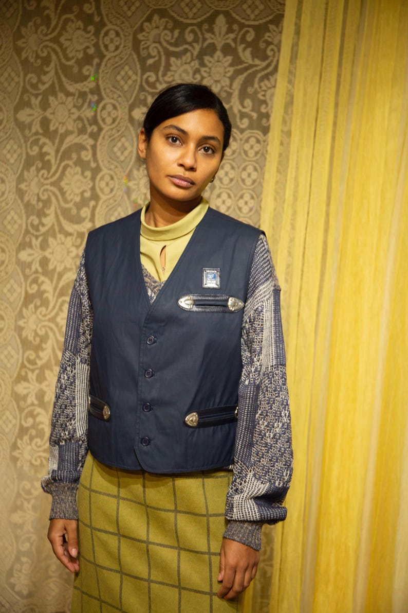 May include: A navy blue waistcoat with silver-toned details and a small square badge. It's worn over a patterned jumper and a pale green roll neck. The outfit is completed with a green plaid skirt.