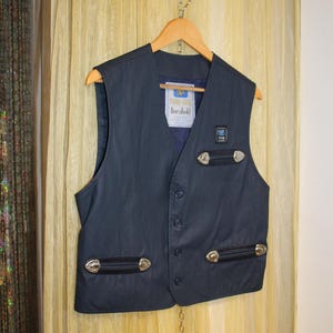May include: A navy blue waistcoat with a V-neck, featuring silver-toned accents and a label that reads "PERRO GOMEZ lineabold." The waistcoat has a button-up front and is hanging on a wooden hanger.