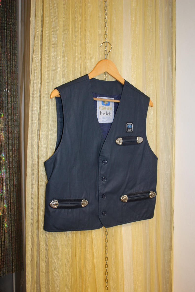 May include: A navy blue waistcoat with a V-neck, featuring silver-toned accents and a label that reads "PERRO GOMEZ lineabold." The waistcoat has a button-up front and is hanging on a wooden hanger.
