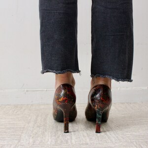 Puede incluir: Un par de zapatos de tac&oacute;n alto con estampado de piel de serpiente en tonos marrones, naranjas y verdes. Los zapatos tienen punta puntiaguda y tac&oacute;n de aguja.