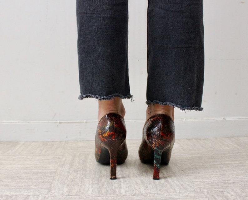 Puede incluir: Un par de zapatos de tac&oacute;n alto con estampado de piel de serpiente en tonos marrones, naranjas y verdes. Los zapatos tienen punta puntiaguda y tac&oacute;n de aguja.