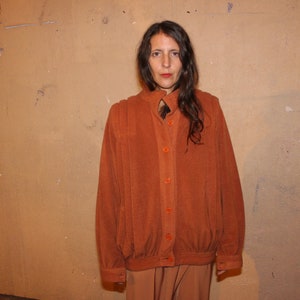 Może przedstawiać: Rdzawo-pomarańczowa kurtka z zapięciem na guziki i wysokim kołnierzem. Kurtka ma lekko powiększony kr&oacute;j i jest zestawiona z pasującymi spodniami. Str&oacute;j jest noszony przez osobę stojącą przed jasnobrązową ścianą.
