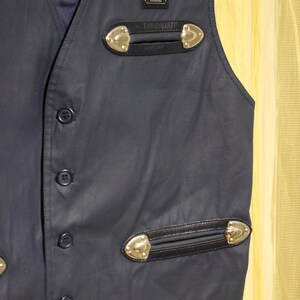 May include: A navy blue waistcoat with decorative gold-coloured accents and black leather trim. The waistcoat has a button-up front and a small label with text. The waistcoat has two decorative pockets with gold-coloured accents.