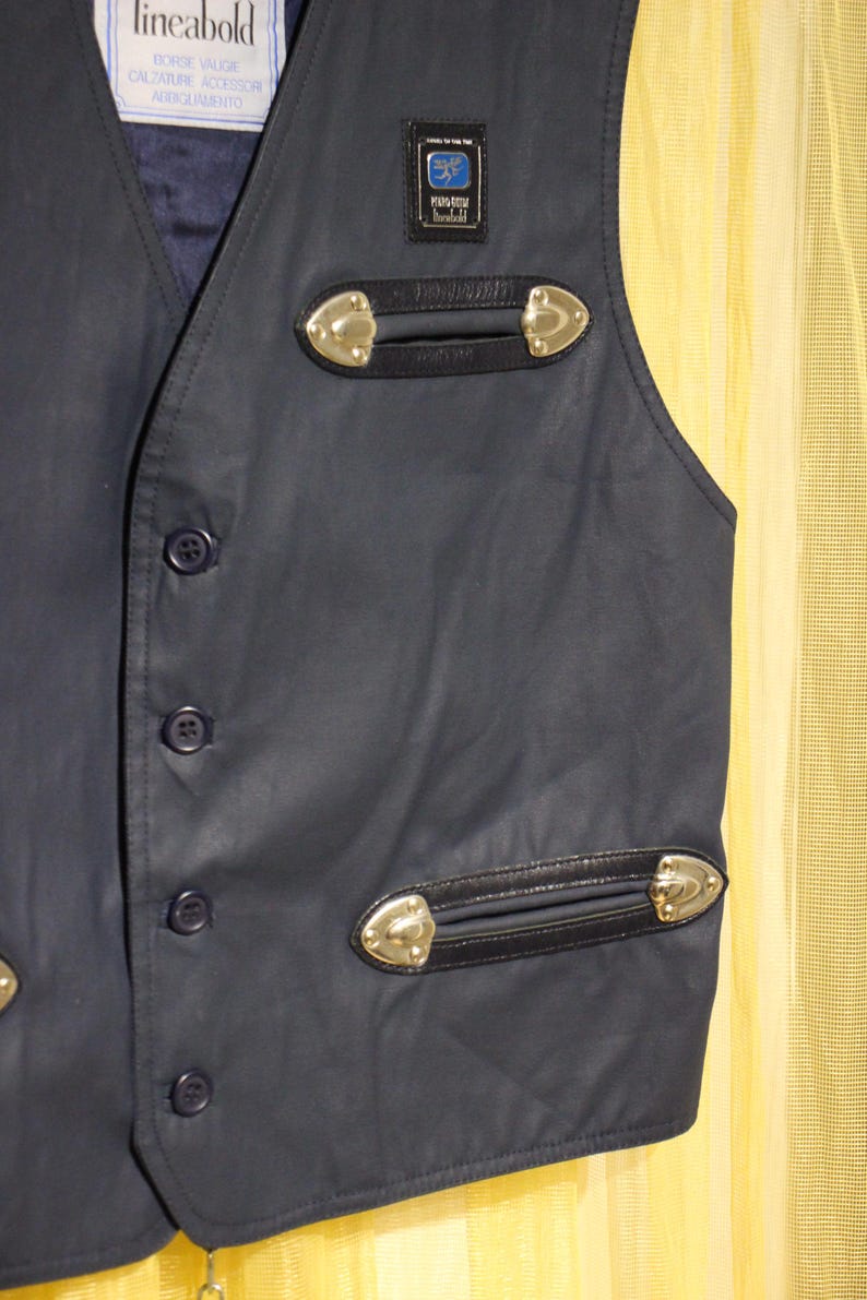 May include: A navy blue waistcoat with decorative gold-coloured accents and black leather trim. The waistcoat has a button-up front and a small label with text. The waistcoat has two decorative pockets with gold-coloured accents.