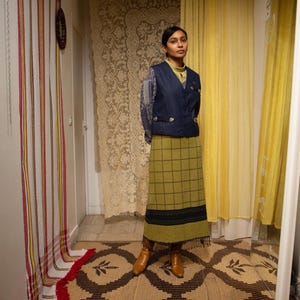 May include: A woman in a navy blue vest, light green top, and olive green plaid skirt. She wears brown boots. The background features a patterned rug, lace curtains, and yellow curtains. The skirt has a fringe.