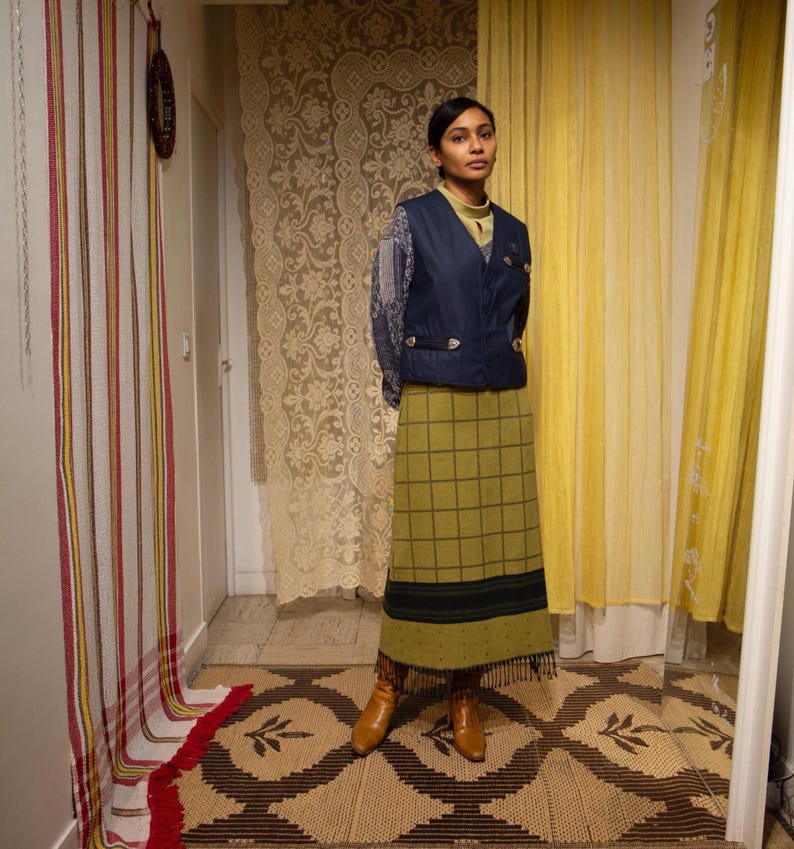 May include: A woman in a navy blue vest, light green top, and olive green plaid skirt. She wears brown boots. The background features a patterned rug, lace curtains, and yellow curtains. The skirt has a fringe.