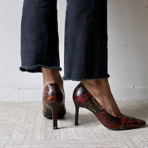 Puede incluir: Un par de zapatos de tac&oacute;n alto con estampado de piel de serpiente en tonos rojos, negros y verdes. Los zapatos tienen punta puntiaguda y tac&oacute;n de aguja.