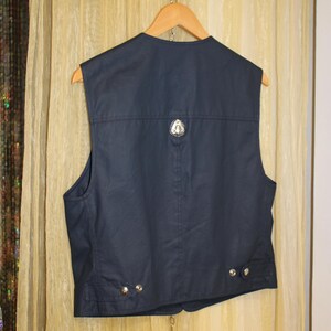 May include: Navy blue vest with a silver emblem on the back and silver buttons. The sleeveless vest hangs on a wooden hanger. The vest is made of a smooth, possibly waxed, fabric.