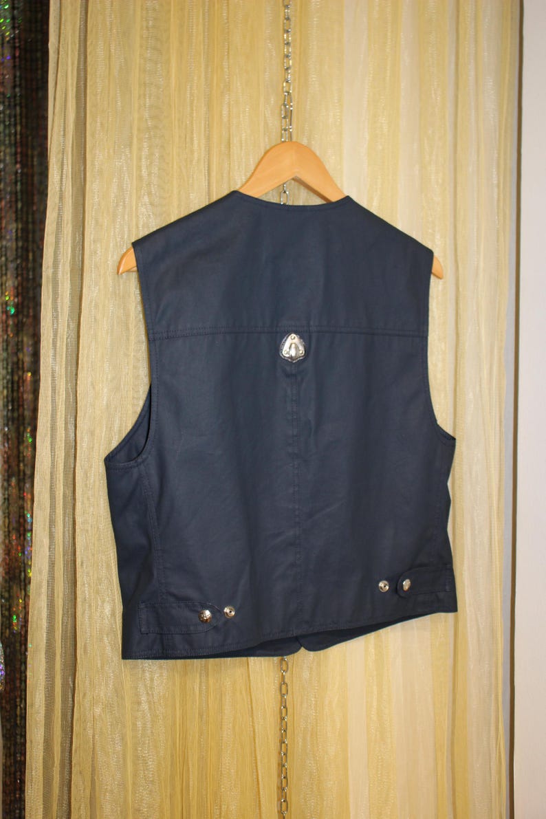 May include: Navy blue vest with a silver emblem on the back and silver buttons. The sleeveless vest hangs on a wooden hanger. The vest is made of a smooth, possibly waxed, fabric.