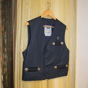 May include: Dark navy vest with silver-coloured embellishments and black leather details. The vest features a button-up front and a label reading "linebold". It is suspended on a wooden hanger.