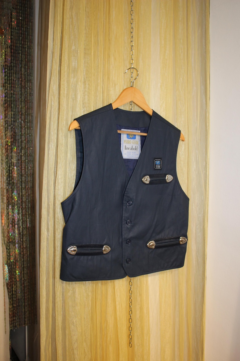 May include: Dark navy vest with silver-coloured embellishments and black leather details. The vest features a button-up front and a label reading "linebold". It is suspended on a wooden hanger.