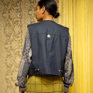 May include: A navy blue vest with a decorative silver button is worn over a patterned sweater. The model is wearing a green plaid skirt. The background features a floral patterned wallpaper and a yellow curtain.