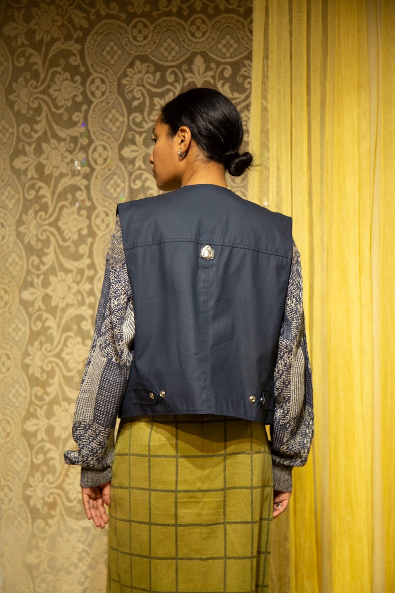 May include: A navy blue vest with a decorative silver button is worn over a patterned sweater. The model is wearing a green plaid skirt. The background features a floral patterned wallpaper and a yellow curtain.