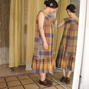 May include: A sleeveless plaid dress in shades of yellow, brown, blue, and orange. The dress has a ruffled hem and is worn with brown leather shoes. The person is standing in front of a mirror, reflecting the dress.