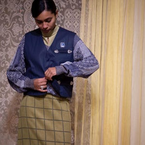 May include: A person wearing a navy blue vest over a patterned sweater and a green plaid skirt. The vest has silver-coloured clasps and a small emblem. The background features a floral wallpaper and a yellow curtain.