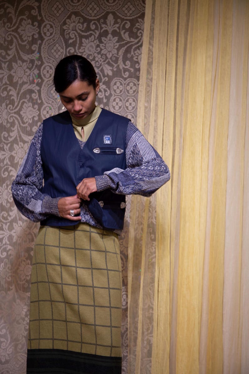 May include: A person wearing a navy blue vest over a patterned sweater and a green plaid skirt. The vest has silver-coloured clasps and a small emblem. The background features a floral wallpaper and a yellow curtain.