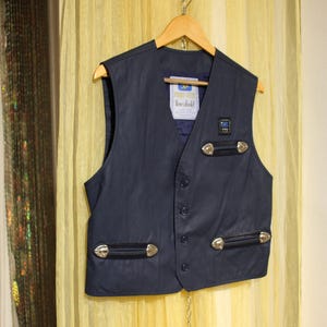 May include: A navy blue waistcoat with silver-coloured embellishments and decorative features. The waistcoat has a button-up front and two pockets with silver-coloured hardware. It hangs on a wooden hanger against a yellow backdrop.