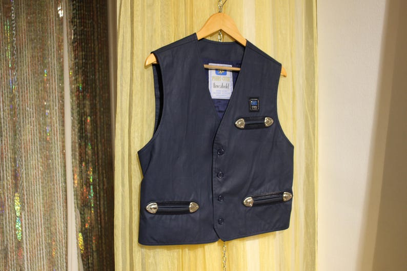 May include: A navy blue waistcoat with silver-coloured embellishments and decorative features. The waistcoat has a button-up front and two pockets with silver-coloured hardware. It hangs on a wooden hanger against a yellow backdrop.
