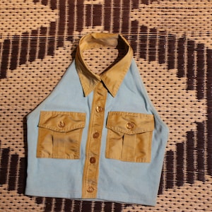 May include: A light blue halter top with a tan collar, button-up front, and two front pockets. The top is designed to resemble a collared shirt. The background is a woven brown and tan pattern.