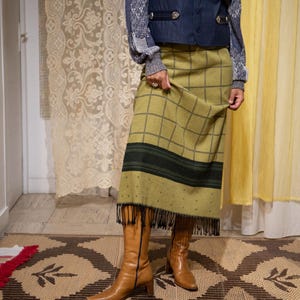 May include: An outfit featuring a navy blue waistcoat, a patterned long-sleeve shirt, and a green plaid skirt with fringe. The outfit is paired with brown leather boots. The skirt has a black stripe and black fringe.