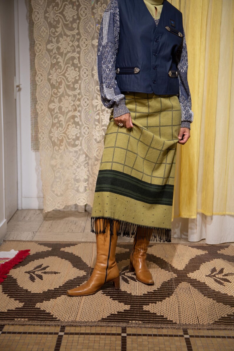 May include: An outfit featuring a navy blue waistcoat, a patterned long-sleeve shirt, and a green plaid skirt with fringe. The outfit is paired with brown leather boots. The skirt has a black stripe and black fringe.