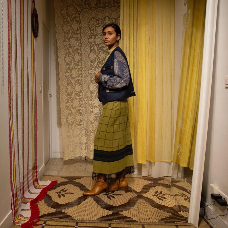 May include: A person wearing a dark blue denim jacket with patchwork sleeves, a long green plaid skirt, and brown boots. The skirt features a black stripe and fringe at the hem. The background includes patterned curtains and a decorative rug.