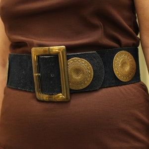 May include: A brown suede belt with a large brass buckle and three decorative round brass accents. The belt is worn over a brown dress.