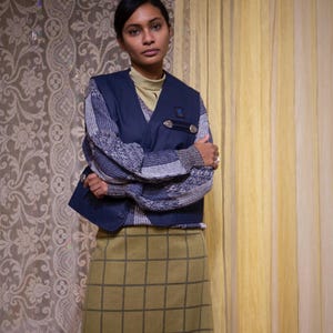May include: A person wearing a navy blue vest over a patterned sweater and a green grid-patterned skirt. The vest has silver button details. The background features patterned wallpaper and yellow curtains.