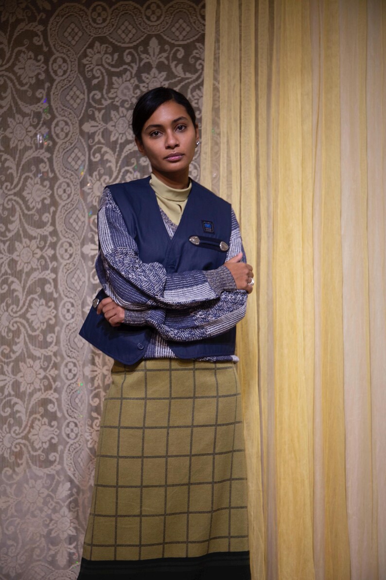 May include: A person wearing a navy blue vest over a patterned sweater and a green grid-patterned skirt. The vest has silver button details. The background features patterned wallpaper and yellow curtains.
