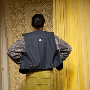 May include: A navy blue vest with a silver button detail is worn over a patterned long-sleeved shirt and a yellow plaid skirt. The person's back is facing the camera, with their hands on their hips.