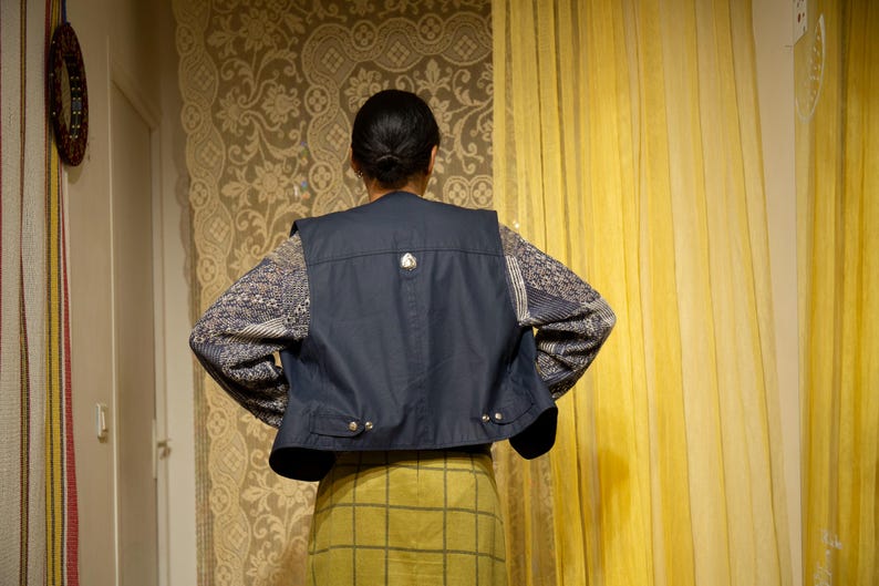 May include: A navy blue vest with a silver button detail is worn over a patterned long-sleeved shirt and a yellow plaid skirt. The person's back is facing the camera, with their hands on their hips.